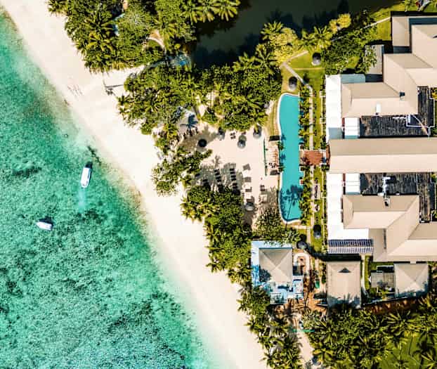 Flygbild över en strand och resort med palmträd, pool och byggnader. (AI-genererad text)