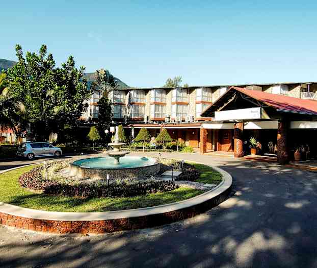 Hotellkomplex med stora fönster, välskött trädgård och en fontän, beläget mot en bakgrund av berg. (AI-genererad text)