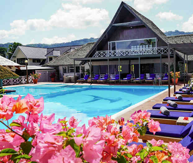 Poolområde med solstolar och blommor, byggnader i bakgrunden. (AI-genererad text)