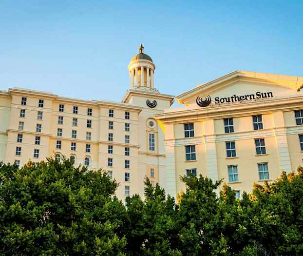 En ljus byggnad med flera våningar och ett torn syns bakom grönskande träd. (AI-genererad text)