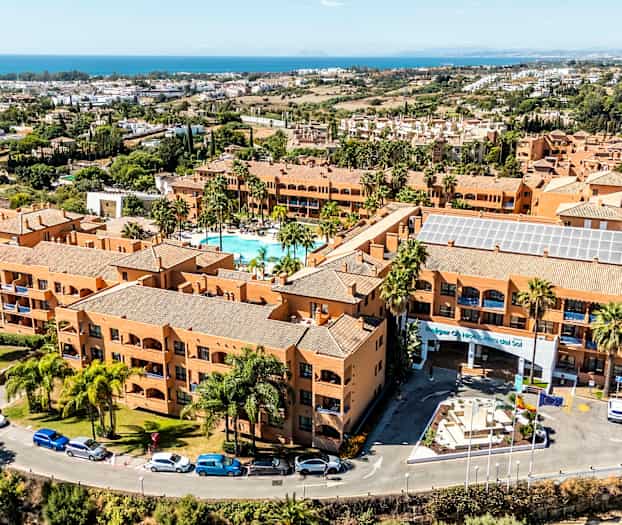 Ett semesterresort med terrakottafärgade byggnader, en pool i centrum och omgivande grönska. Havet syns i bakgrunden. (AI-genererad text)