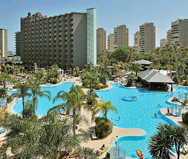 Ett hotellområde med stor pool och solstolar, omgiven av palmer och höghus. (AI-genererad text)