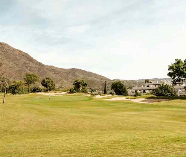 En golfbana omgiven av grön vegetation, med byggnader och berg i bakgrunden. (AI-genererad text)