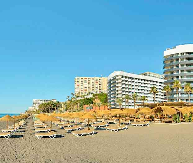 En strand med solstolar och parasoller, omgiven av flera hotellbyggnader. (AI-genererad text)