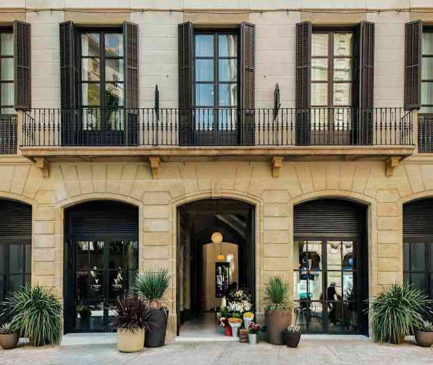 Bild av fasaden på en byggnad med flera höga fönster och ett ingångsparti med dekorationer av blomkrukor. (AI-genererad text)