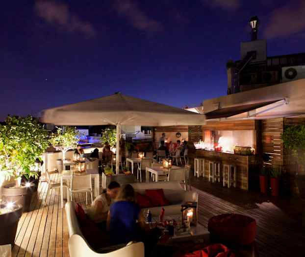 En takterrass med sittgrupper, bord, parasoller och grönska, upplyst i kvällsmiljö. (AI-genererad text)