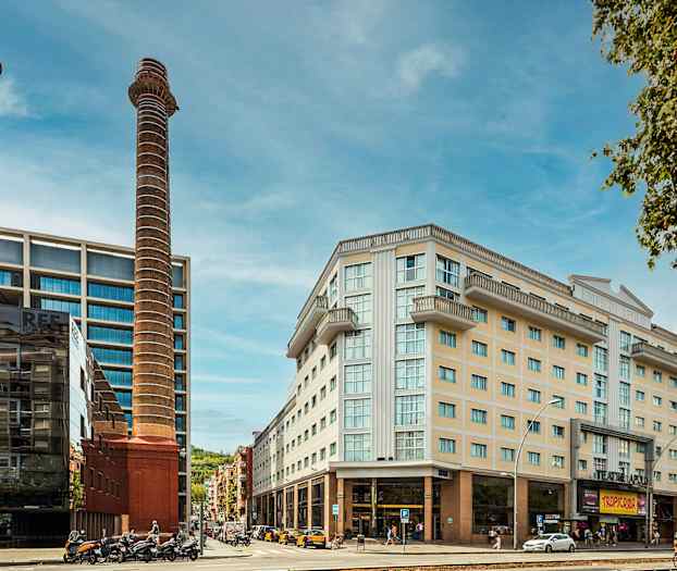 Bild av en stadsmiljö med moderna byggnader, två höga skorstenar och några människor och fordon i förgrunden. (AI-genererad text)