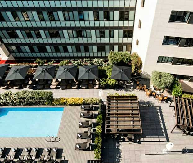 Flygbild över en pool med solstolar, parasoller och omgivande byggnader. (AI-genererad text)