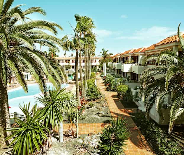 Bild av en semesteranläggning med palmer, trädgårdar och en pool. Byggnader med terrakottafärgade tak syns i bakgrunden. (AI-genererad text)