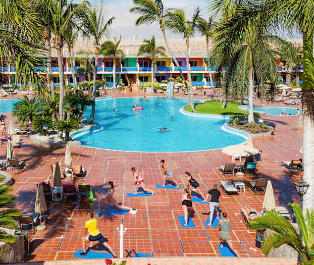 Vid ett stort hotell med färgglada byggnader utövar människor utomhusyoga på mattor bredvid en pool. (AI-genererad text)