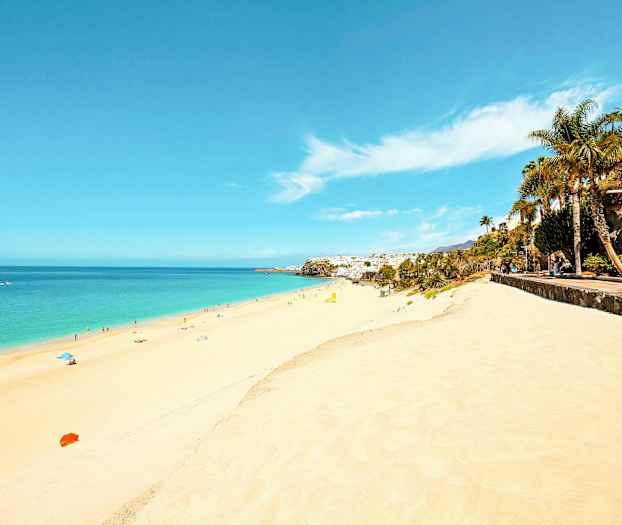 En lång sandstrand med klart blått vatten och människor som solar och badar. Längs sidan finns en promenadväg med palmer och grönska. (AI-genererad text)