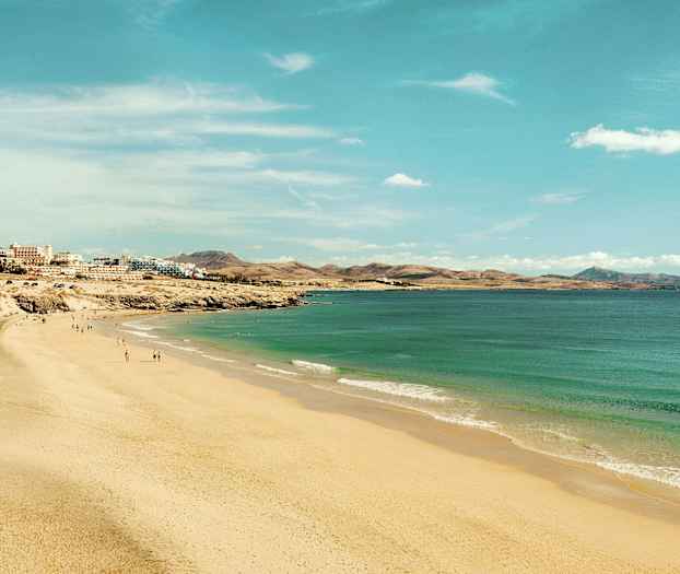 En bred sandstrand som sträcker sig längs en kustlinje med turkost vatten och byggnader i bakgrunden. (AI-genererad text)