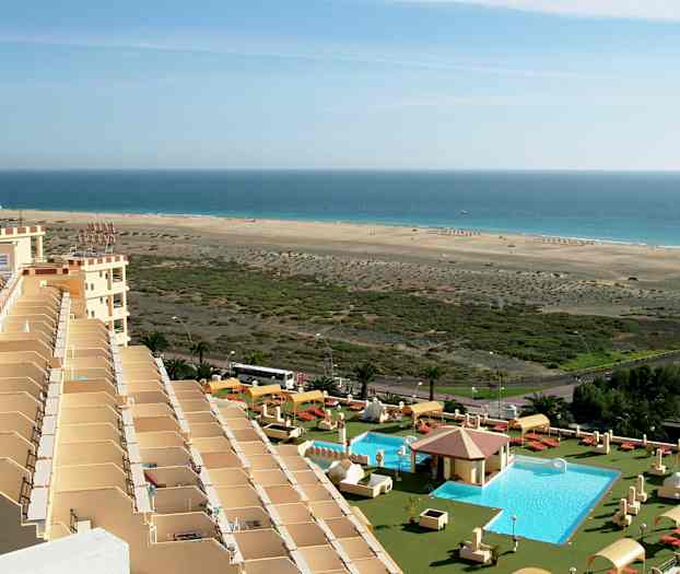 Bild av ett hotellområde med en pool, solstolar och parasoller i förgrunden, och en lång sandstrand vid havet i bakgrunden. (AI-genererad text)