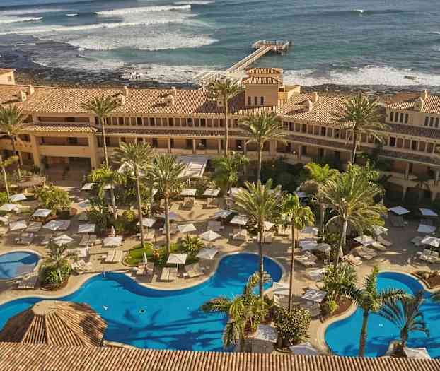 Hotellområde med pooler, solstolar och parasoller omgivet av palmer. I bakgrunden syns havet med vågor och en brygga. (AI-genererad text)