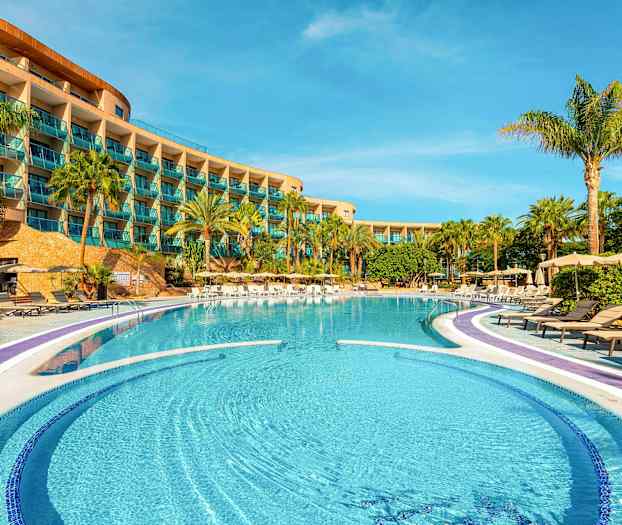 Hotellets poolområde med solstolar och palmer framför en byggnad med balkonger. (AI-genererad text)