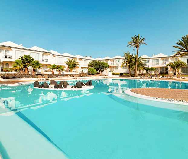 En bild på en hotellpool med klarblått vatten, omgärdad av palmträd och vita byggnader i bakgrunden. (AI-genererad text)