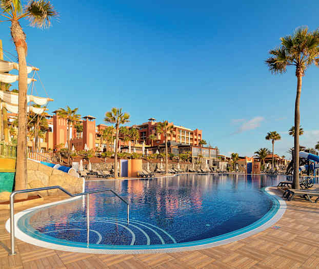 En utomhuspool med soldäck, omgiven av palmträd och med en byggnad i bakgrunden. I förgrunden syns ett skepp som prydnad. (AI-genererad text)