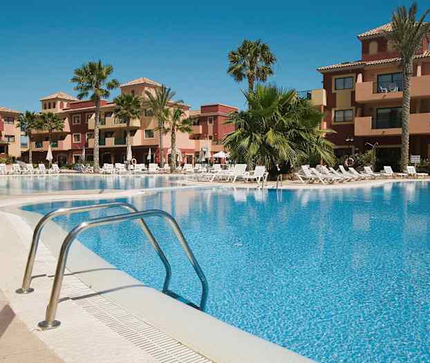 Ett semesterkomplex med en utomhuspool omgiven av solstolar och palmer, samt byggnader i bakgrunden. (AI-genererad text)