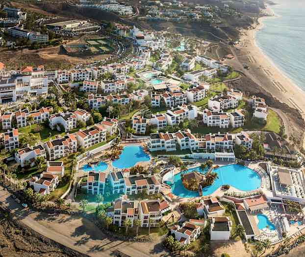 Flygvy över en resort med vita byggnader och röda tak, samt stora pooler och trädgårdar nära stranden. (AI-genererad text)