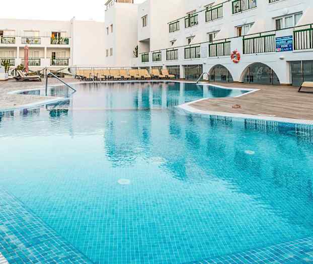 Utomhuspool vid en semesteranläggning med solstolar och vita byggnader i bakgrunden. (AI-genererad text)