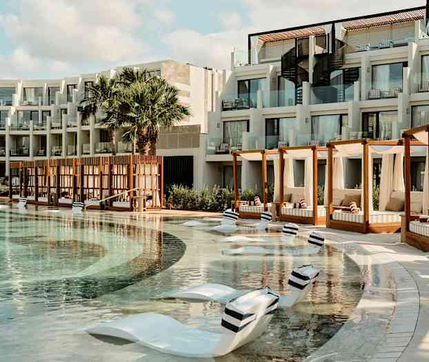 En hotellpool med solstolar i poolen och cabanas på sidan. Byggnaden har balkonger med glasräcken. (AI-genererad text)