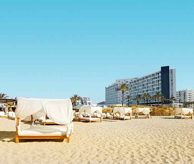 Strand med sängar med vita draperingar, i bakgrunden syns en modern byggnad. (AI-genererad text)