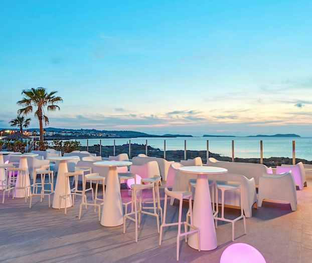 En terrass med vita bord och stolar, omgiven av glaspaneler, med utsikt över havet och en palmliknande växt i bakgrunden. (AI-genererad text)