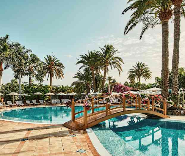 Bild på en utomhuspool med solstolar, parasoller och palmer i bakgrunden. En liten dekorativ bro sträcker sig över vattnet. (AI-genererad text)