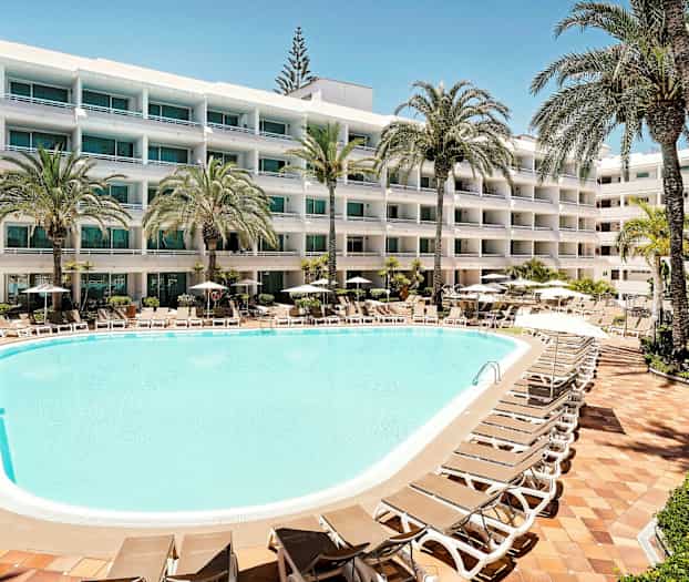 En hotellpool med solstolar och parasoller omgiven av lummiga palmer, samt en flerfaldig byggnad med balkonger i bakgrunden. (AI-genererad text)