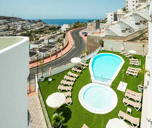 Utomhuspool med solstolar på grön gräsmatta och utsikt över närliggande byggnader och havet. (AI-genererad text)