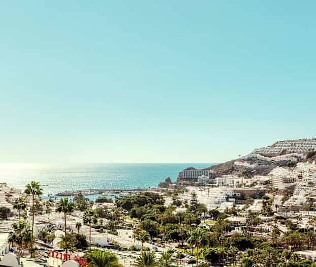 Vy över en kuststad med vita byggnader och palmer framför ett glittrande hav. (AI-genererad text)