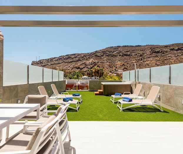 En utomhusterrass med solstolar på konstgräs och en jacuzzi, omgiven av glasbarriär och berg i bakgrunden. (AI-genererad text)
