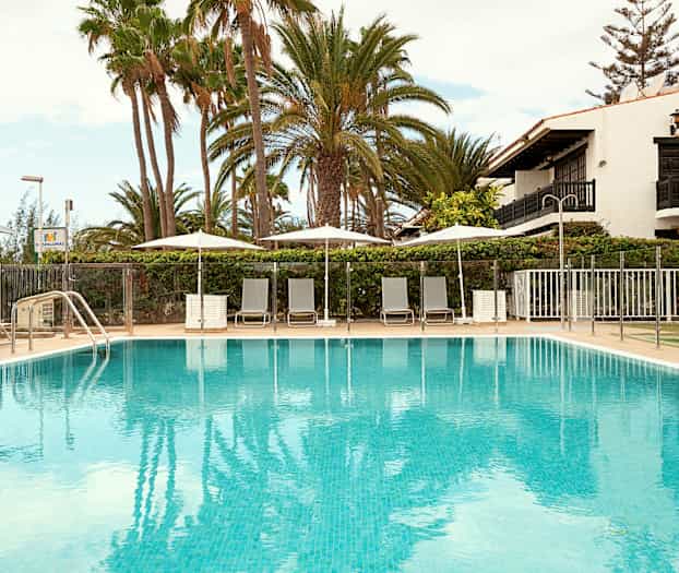 Bild på en utomhuspool omgiven av solstolar och parasoller, med palmträd och en byggnad i bakgrunden. (AI-genererad text)