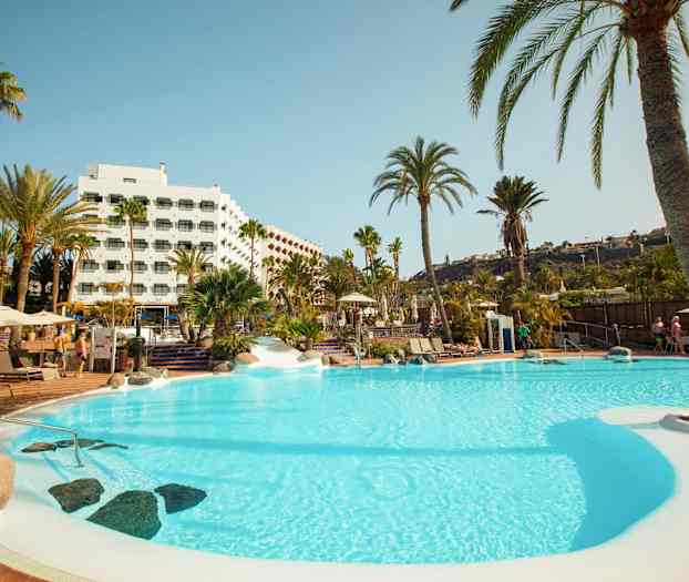 En swimmingpool omgiven av palmträd och ett hotell i bakgrunden. (AI-genererad text)