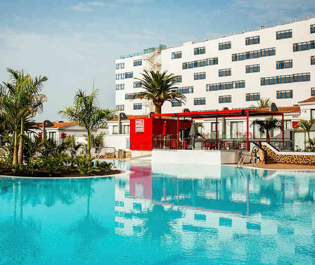 Ett hotellområde med en stor pool, omgiven av palmer och byggnader med vita fasader. (AI-genererad text)