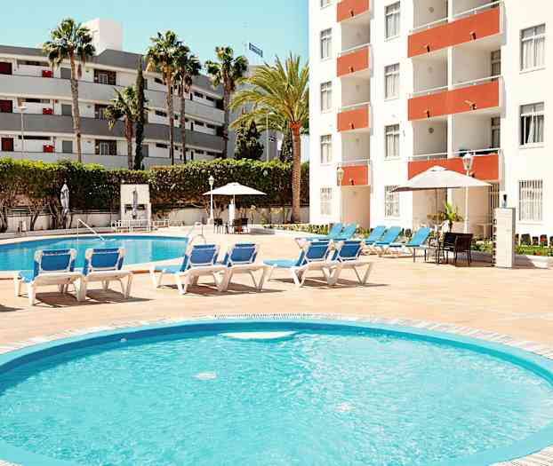 En utomhuspool omgiven av solstolar, med en byggnad i bakgrunden. (AI-genererad text)