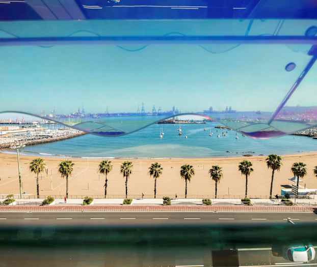 Utsikt över en sandstrand med palmer, en väg i förgrunden och en marina samt en hamn i bakgrunden. (AI-genererad text)