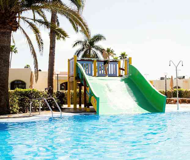 En vattenrutschkana vid en pool, omgiven av palmer och byggnader. (AI-genererad text)