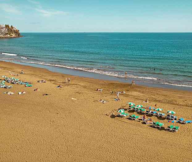 Bilden visar en sandstrand vid havet med solstolar och parasoller samt människor som njuter av stranden. (AI-genererad text)