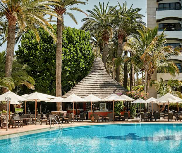 Uteplats med parasoller framför en pool och en byggnad med palmer i bakgrunden. (AI-genererad text)