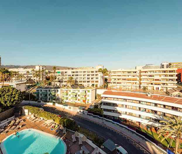 Vy över ett hotellområde med pool, omgiven av byggnader under en klar himmel. (AI-genererad text)