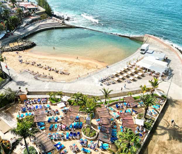 Flygbild av en strand med solstolar och parasoller, omgiven av palmer och en gångväg. (AI-genererad text)