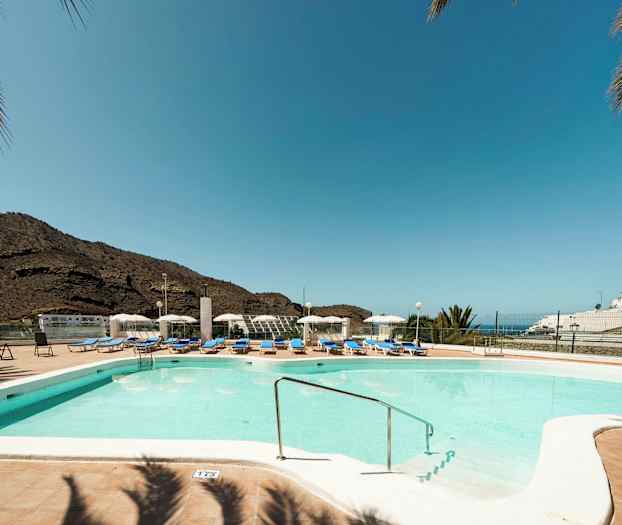 Utomhuspool med solstolar och parasoll, berg i bakgrunden och klarblå himmel. (AI-genererad text)