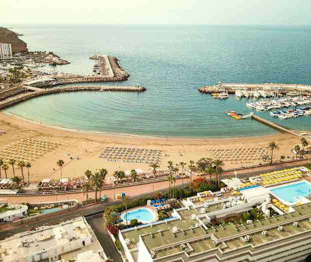 Flygfoto över en strand med parasoller och solstolar, en omgivande marina med båtar, och närliggande byggnader. (AI-genererad text)