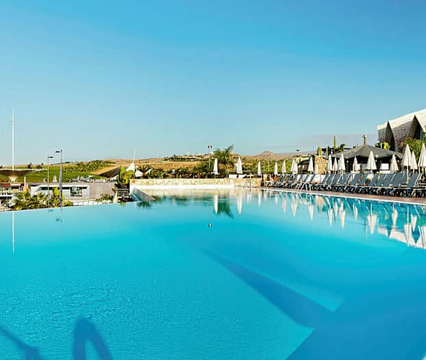 Stor pool utomhus med solstolar och parasoller i bakgrunden, omgiven av grönska och byggnader. (AI-genererad text)