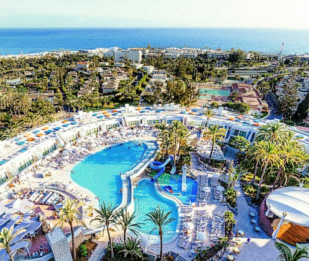 Flygbild över en stor resort med poolområde och omgivet av palmer. (AI-genererad text)