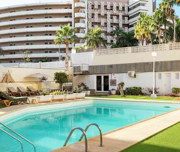 En utomhuspool omgiven av solstolar, grönska och en byggnad i bakgrunden. (AI-genererad text)