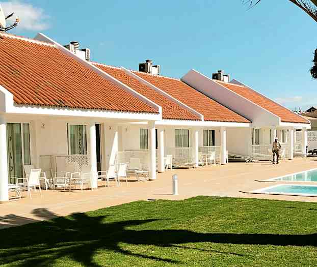 Vita bungalows med orangea tak och en pool framför. En gräsmatta i förgrunden. (AI-genererad text)