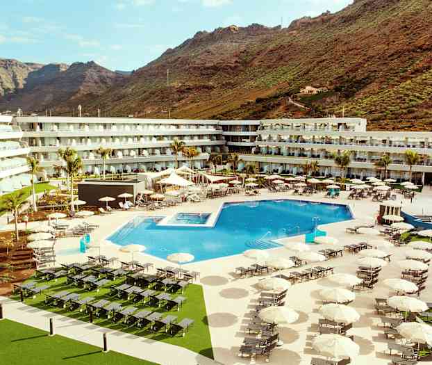 Ett hotellområde med en stor utomhuspool, solstolar och parasoller, omgivet av byggnader och berg i bakgrunden. (AI-genererad text)