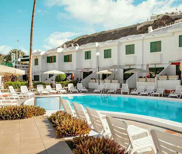 Poolområde med solstolar och parasoller framför ett vitt hus med gröna fönsterluckor. (AI-genererad text)